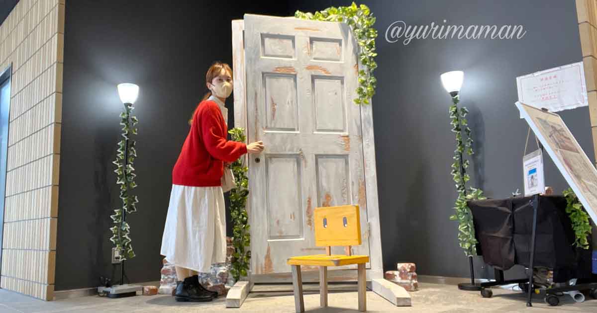 Person in a red sweater and white skirt, wearing a mask, leans on a tall weathered door in a gallery-like space with ivy and lamps nearby.