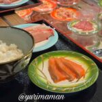 Bowl of steamed white rice on a black tray with a small plate of salmon sashimi nearby, hand resting at left.