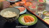 Bowl of steamed white rice on a black tray with a small plate of salmon sashimi nearby, hand resting at left.