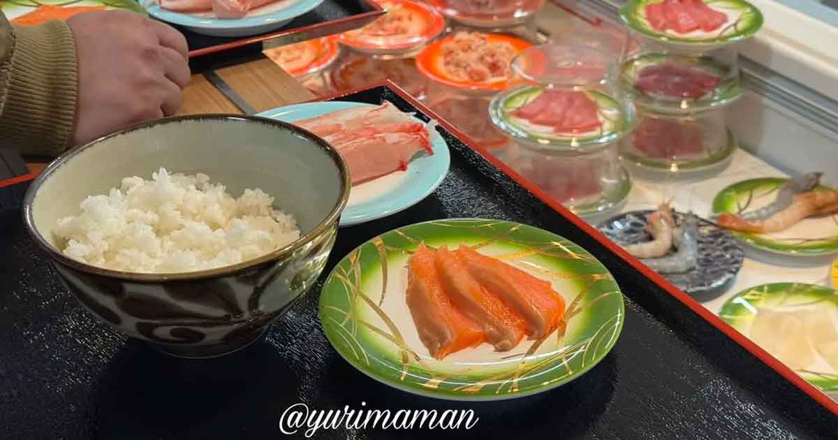 Bowl of steamed white rice on a black tray with a small plate of salmon sashimi nearby, hand resting at left.