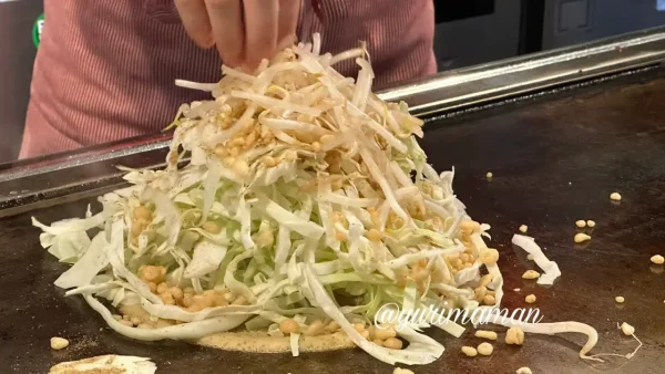 鉄板の上で広島風お好み焼きを調理する様子、山盛りキャベツと天かす