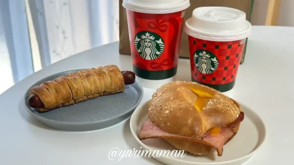 スターバックスのホリデーカップと紙袋、ベーグルサンドとソーセージパイのテイクアウトパッケージを並べた写真。