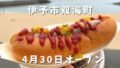 Hot dog with ketchup and mustard on a bun held at the beach; text announces opening on April 30 in Iyo City Futami Town (Futami-cho).