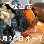 Rice balls wrapped in seaweed on a wooden board, with Japanese text announcing Matsuyama City and an April 25 opening.