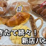 Assorted freshly baked breads in baskets and bags on a table, with bold Japanese text advertising a new bakery.