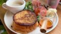 Brunch plate with a stack of French toast, syrup pitcher, coffee, greens, tomato, cheese, and ham on a wooden table.