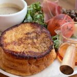 Brunch plate with a stack of French toast, syrup pitcher, coffee, greens, tomato, cheese, and ham on a wooden table.