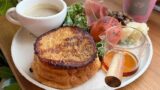 Brunch plate with a stack of French toast, syrup pitcher, coffee, greens, tomato, cheese, and ham on a wooden table.