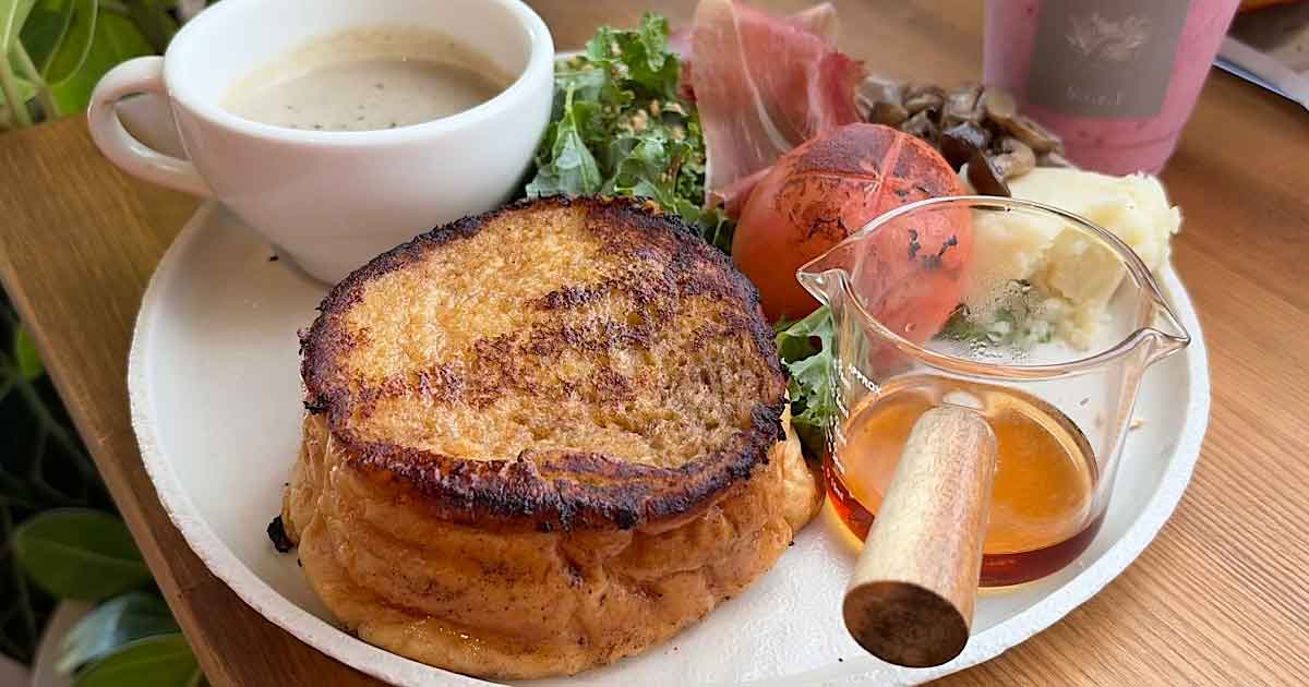 Brunch plate with a stack of French toast, syrup pitcher, coffee, greens, tomato, cheese, and ham on a wooden table.