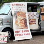 Mobile sandwich van with a large banner advertising HOT SAND and SWEET SAND, and a sandwich-board sign in front displaying the menu
