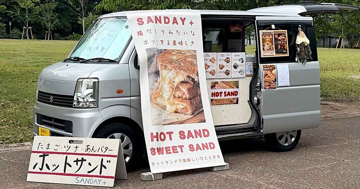 Mobile sandwich van with a large banner advertising HOT SAND and SWEET SAND, and a sandwich-board sign in front displaying the menu