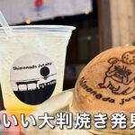 Plastic cup with straw and pale yellow beverage next to a round dorayaki pancake with a printed cartoon image on a wooden counter