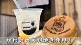 Plastic cup with straw and pale yellow beverage next to a round dorayaki pancake with a printed cartoon image on a wooden counter