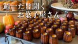 Text in Japanese reads '週一だけ開くお菓子屋さん' over a display of canelé pastries on a tray with pumpkins and fruit in the background.