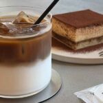Iced coffee with ice cubes and milk layers in a glass with a straw, beside a tiramisu slice on a plate with a fork nearby.