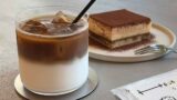 Iced coffee with ice cubes and milk layers in a glass with a straw, beside a tiramisu slice on a plate with a fork nearby.