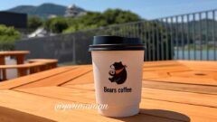 Disposable coffee cup with Bears coffee logo on a wooden outdoor table, bright sunny day with railing and hills in the background.