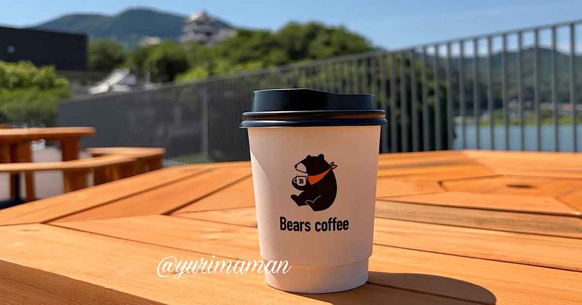 Disposable coffee cup with Bears coffee logo on a wooden outdoor table, bright sunny day with railing and hills in the background.