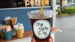 Iced coffee in a clear cup with a round logo showing a fish and the text がま cafe, held outdoors near a cafe counter.