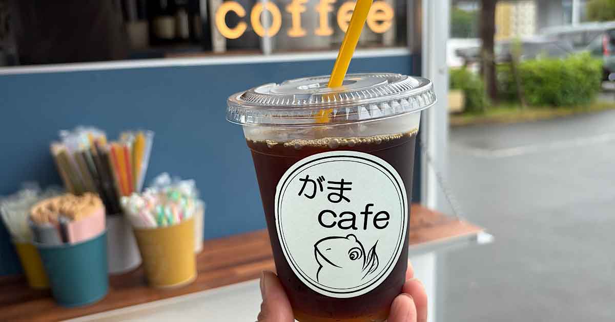 Iced coffee in a clear cup with a round logo showing a fish and the text がま cafe, held outdoors near a cafe counter.
