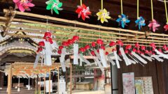Shinto shrine courtyard with rows of red ornaments and white paper strips hanging from ropes under a wooden beam; colorful pinwheels above.