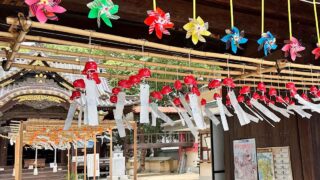 松山市・三津厳島神社の夏詣で設置された風鈴スポット - ゆりママんブログ Shinto shrine courtyard with rows of red ornaments and white paper strips hanging from ropes under a wooden beam; colorful pinwheels above.